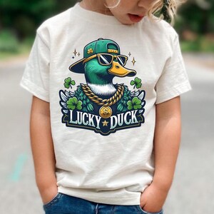 K&ouml;nnte beinhalten: Ein wei&szlig;es T-Shirt mit einer Cartoon-Illustration eines Entchens, das eine gr&uuml;n-goldene Baseballkappe, Sonnenbrille und eine goldene Kette tr&auml;gt. Das Entchen ist von gr&uuml;nen Kleebl&auml;ttern umgeben und der Text "Lucky Duck" ist unter dem Entchen gedruckt.