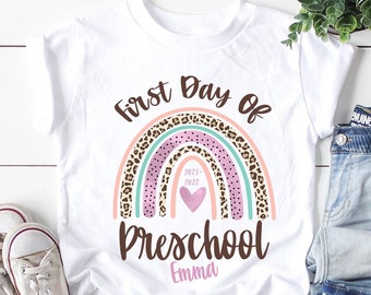 May include: White t-shirt with a rainbow design featuring a pink heart and the text "First Day of Preschool" with the name "Emma" underneath.