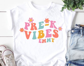 May include: White t-shirt with the text "PRE-K VIBES" in retro colors and floral accents. The name "EMMY" is below the text. The shirt is paired with light wash denim jeans and white sneakers. A small potted plant is also in the image.