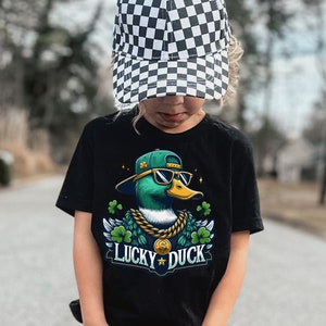 K&ouml;nnte beinhalten: Ein Kind tr&auml;gt ein schwarzes T-Shirt mit einer Comic-Ente, die eine Sonnenbrille und eine goldene Kette tr&auml;gt. Die Ente ist von gr&uuml;nen Kleebl&auml;ttern umgeben und der Text "Lucky Duck" ist zu sehen. Das Kind tr&auml;gt auch eine schwarz-wei&szlig; karierte Baseballkappe und eine blaue Jeans.