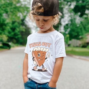 May include: A young child wearing a white t-shirt with a cartoon pie graphic and the text "Piece Out" in orange letters. The child is wearing blue jeans and a black baseball cap. The child is standing on a paved road with a brown dog sitting in front of them.