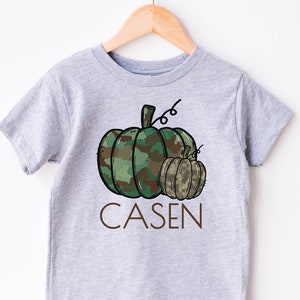 May include: A gray short-sleeved t-shirt with a camouflage pumpkin and a smaller pumpkin. The text "CASEN" is printed below the pumpkins.