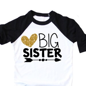 black sleeve raglan shirt with black wording big sister and a arrow with a gold glitter heart