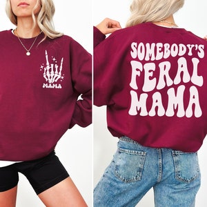 May include: Burgundy sweatshirt with white graphic text and design. The front features a skeleton hand making a rock-and-roll gesture with the word "MAMA" below. The back of the sweatshirt says "SOMEBODY'S FERAL MAMA" in a retro font.