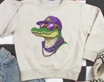 May include: A light gray sweatshirt with a cartoon alligator wearing sunglasses and a purple baseball cap. The alligator is wearing a purple and gold beaded necklace.