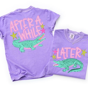 May include: Two purple t-shirts with alligator graphics. One shirt has the text "AFTER A WHILE" and the other says "LATER". The alligators are green with pink and green accents. The shirts have rolled sleeves.
