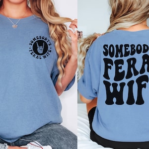 May include: A light blue t-shirt with the text "Somebody's Feral Wife" in black on the back. The front of the shirt has a black circle with the text "Somebody's Feral Wife" and a rock and roll hand symbol.