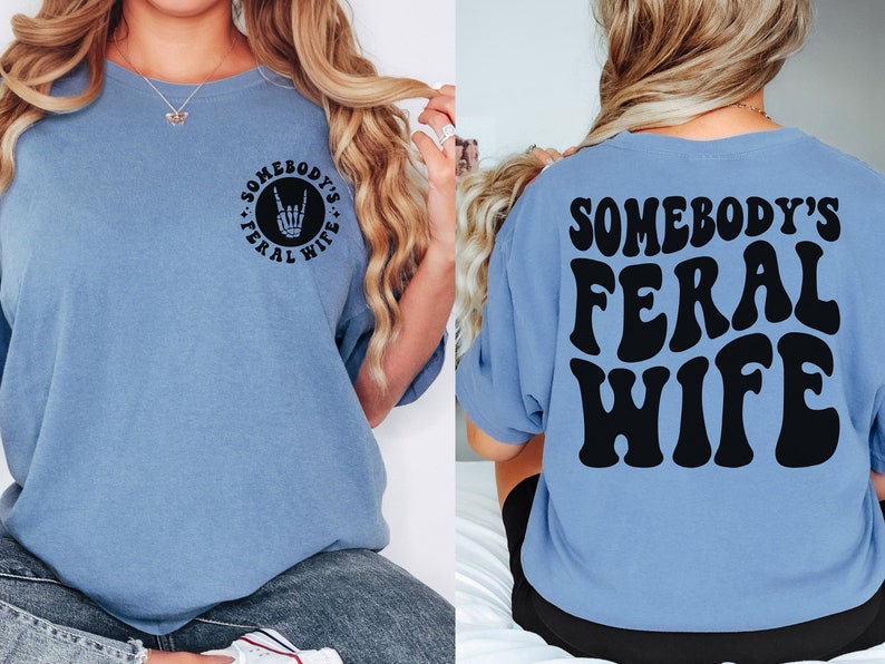 May include: A light blue t-shirt with the text "Somebody's Feral Wife" in black on the back. The front of the shirt has a black circle with the text "Somebody's Feral Wife" and a rock and roll hand symbol.