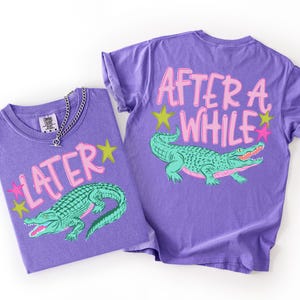 May include: Two purple t-shirts with matching designs. One shirt displays the word "LATER" with a green alligator graphic and star accents. The other shirt has the words "AFTER A WHILE" and a similar alligator design.