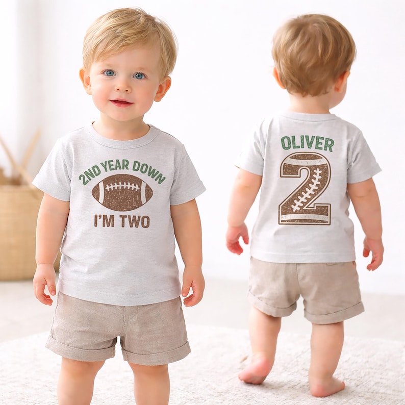 K&ouml;nnte beinhalten: Hellgraues T-Shirt mit einem Football-Motiv und dem Text "2ND YEAR DOWN I'M TWO". Auf der R&uuml;ckseite des Shirts stehen der Name "Oliver" und die Zahl "2" in einem Football-Design. Das Kind tr&auml;gt eine khakifarbene Shorts.