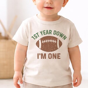 K&ouml;nnte beinhalten: Ein Baby in einem beigen T-Shirt und khakifarbener Shorts. Das T-Shirt hat den Text "1ST YEAR DOWN" &uuml;ber einer braunen Football-Grafik und "I'M ONE" darunter. Das Baby hat hellbraunes Haar und blaue Augen.