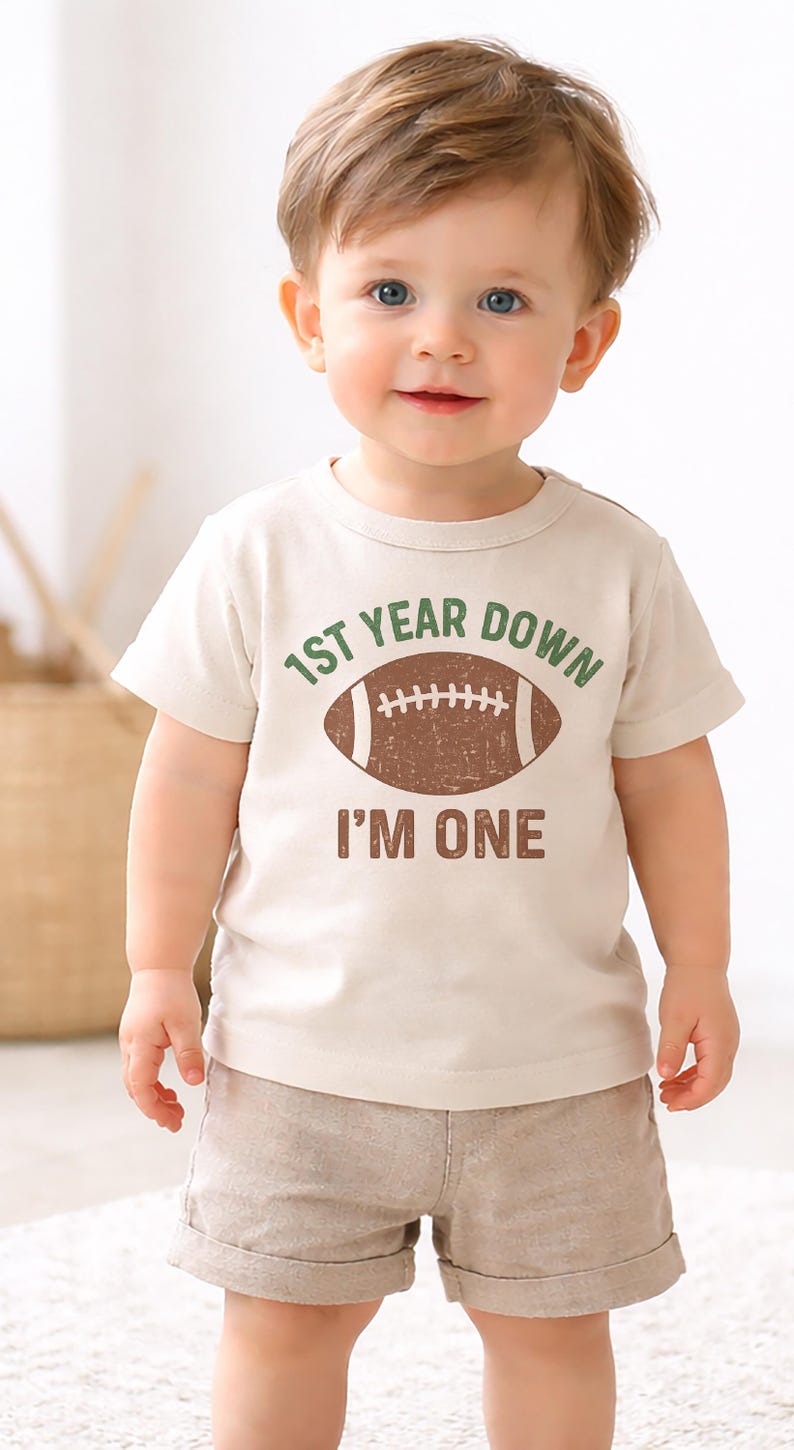 K&ouml;nnte beinhalten: Ein Baby in einem beigen T-Shirt und khakifarbener Shorts. Das T-Shirt hat den Text "1ST YEAR DOWN" &uuml;ber einer braunen Football-Grafik und "I'M ONE" darunter. Das Baby hat hellbraunes Haar und blaue Augen.