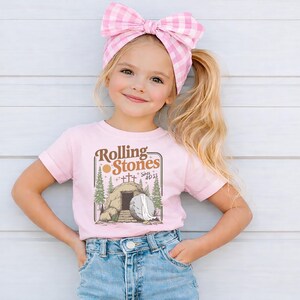 May include: A young girl wearing a pink t-shirt with the text "Rolling Stones" and an illustration of a tomb. She is also wearing blue denim shorts and a pink gingham bow in her hair.