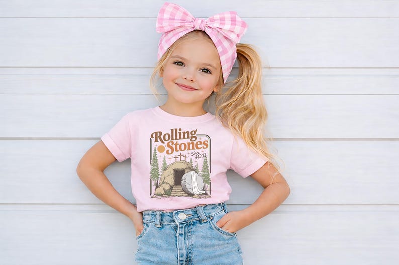 May include: A young girl wearing a pink t-shirt with the text "Rolling Stones" and an illustration of a tomb. She is also wearing blue denim shorts and a pink gingham bow in her hair.