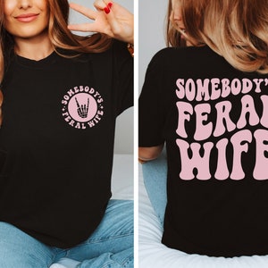 May include: Black t-shirt with a pink graphic on the front that says "Somebody's Feral Wife" with a rock and roll hand symbol. The back of the shirt has a larger pink graphic that says "Somebody's Feral Wife".