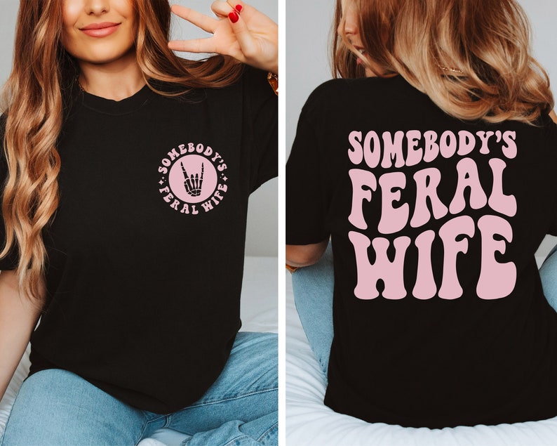 May include: Black t-shirt with a pink graphic on the front that says "Somebody's Feral Wife" with a rock and roll hand symbol. The back of the shirt has a larger pink graphic that says "Somebody's Feral Wife".