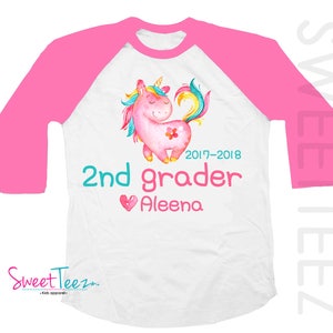 Pode incluir: Camiseta raglan branca e rosa com um desenho de unic&oacute;rnio em aquarela. A camiseta tem o texto "2nd grader Aleena" e os anos "2017-2018". A marca "Sweet Teez" tamb&eacute;m &eacute; vis&iacute;vel.