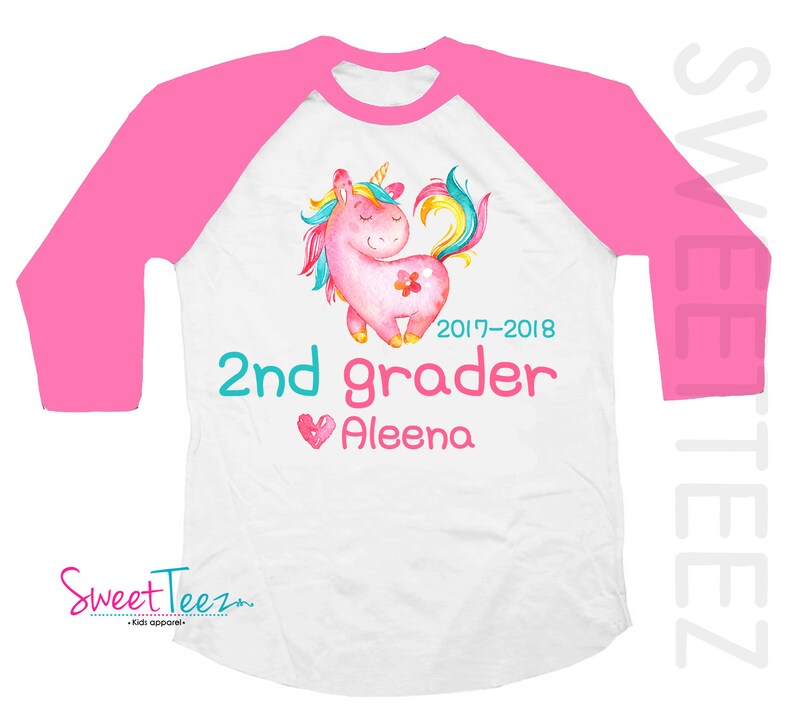 Pode incluir: Camiseta raglan branca e rosa com um desenho de unic&oacute;rnio em aquarela. A camiseta tem o texto "2nd grader Aleena" e os anos "2017-2018". A marca "Sweet Teez" tamb&eacute;m &eacute; vis&iacute;vel.