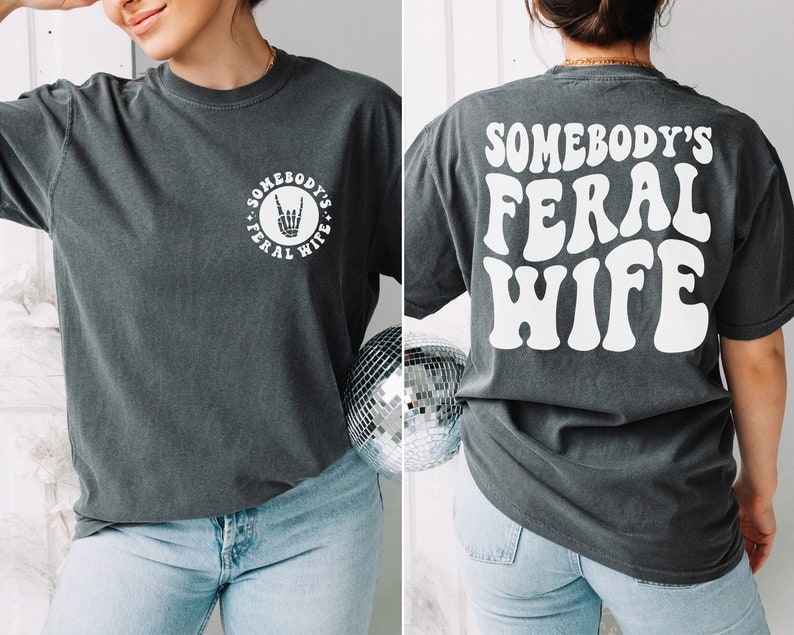 woman wearing a pepper gray comfort colors shirt with the white wording on the back somebody's feral wife and a small pocket skeleton hand on the front