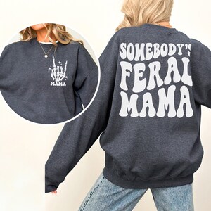 May include: Dark gray sweatshirt with white text and graphics. The front left chest features a white skeleton hand graphic with the word "MAMA" below. The back of the sweatshirt reads "SOMEBODY'S FERAL MAMA" in a retro font.