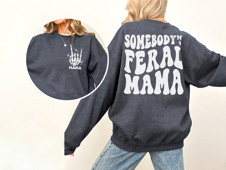 May include: Dark gray sweatshirt with white text and graphics. The front left chest features a white skeleton hand graphic with the word "MAMA" below. The back of the sweatshirt reads "SOMEBODY'S FERAL MAMA" in a retro font.