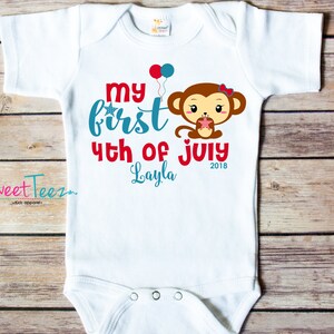 May include: White baby onesie with the text "My First 4th of July Layla 2018" in red and teal. A cartoon monkey holding a cupcake is featured on the front, along with red and blue balloons. The brand "Sweet Teez" is printed on the left side.
