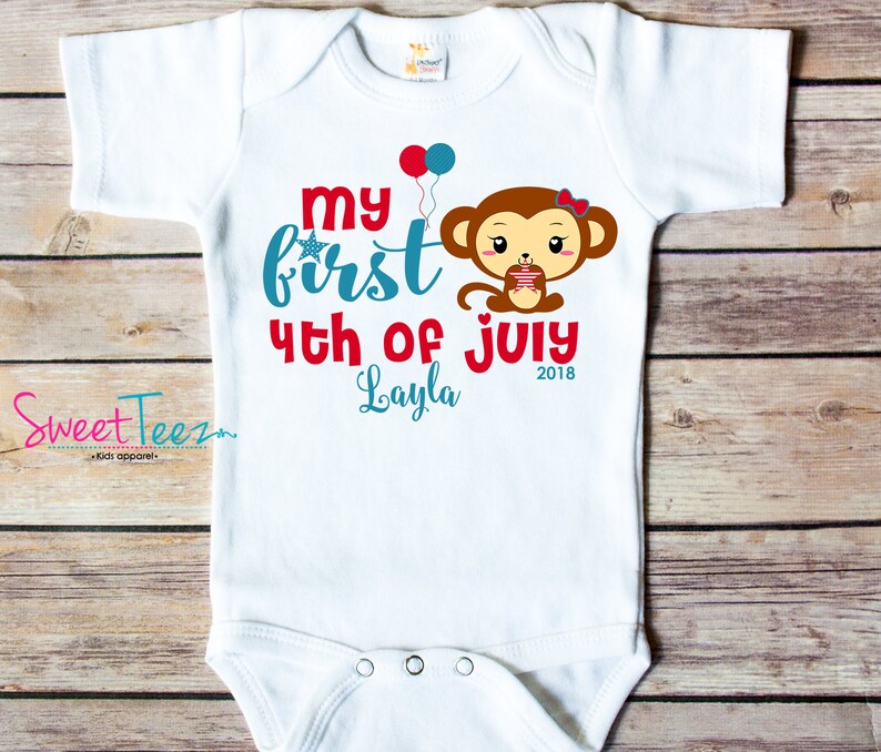 May include: White baby onesie with the text "My First 4th of July Layla 2018" in red and teal. A cartoon monkey holding a cupcake is featured on the front, along with red and blue balloons. The brand "Sweet Teez" is printed on the left side.