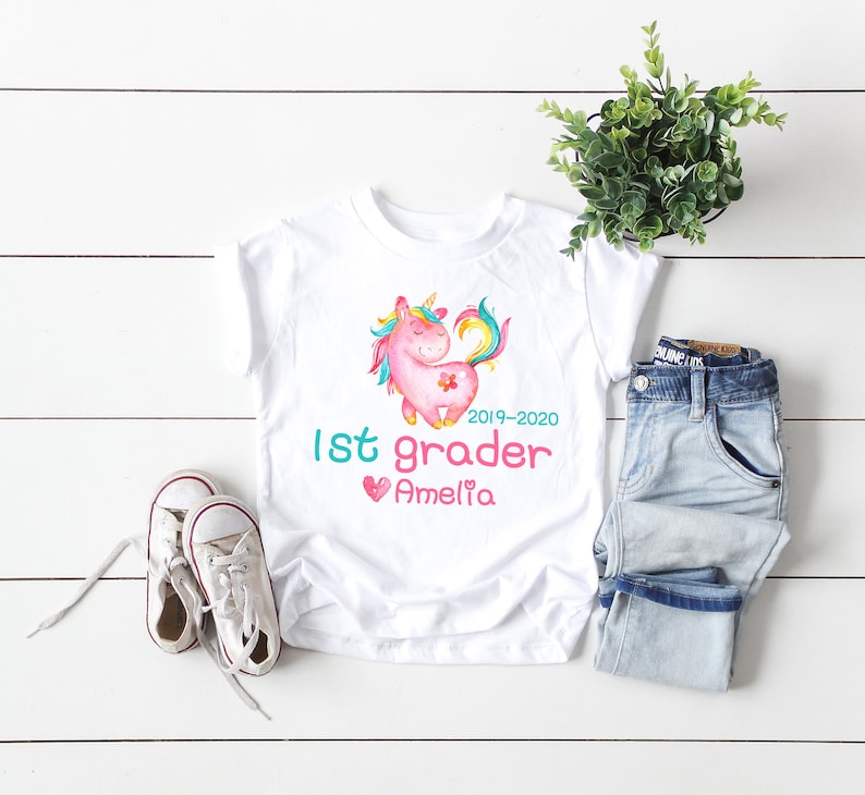 Pode incluir: T-shirt branco de manga curta com um desenho de unic&oacute;rnio em aquarela nas cores rosa, amarelo e azul. A camisa tem o texto "1st grader 2019-2020" e um nome. T&ecirc;nis brancos e jeans azuis ao lado.