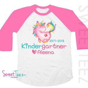 Pode incluir: Camiseta branca e rosa estilo raglan com um design de unic&oacute;rnio em aquarela. O unic&oacute;rnio &eacute; rosa com uma crina e cauda coloridas. A camiseta tem o texto "Kindergartner Aleena" e o ano "2017-2018".