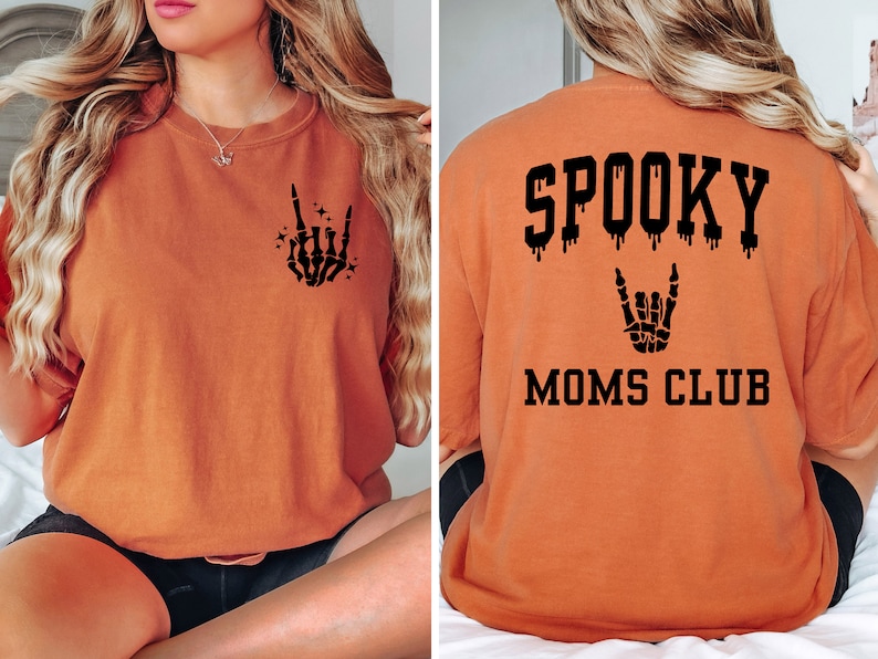 Pu&ograve; includere: Maglietta arancione con una grafica di mano scheletrica nera sul davanti e il testo "SPOOKY MOMS CLUB" con un effetto gocciolante e una grafica di mano scheletrica sul retro.