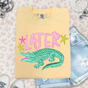 May include: A light yellow t-shirt with the word "LATER" in pink, outlined in a darker pink, and a green alligator graphic. The shirt also features pink and green star graphics. The shirt is paired with denim shorts.
