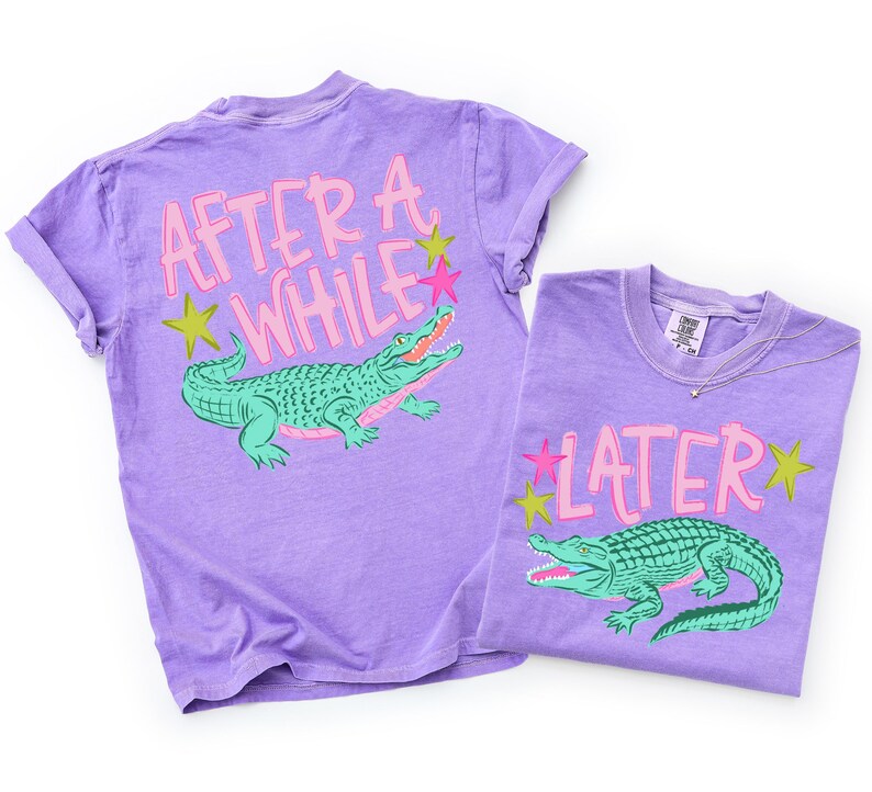 May include: Two purple t-shirts with alligator graphics. One shirt has the text "AFTER A WHILE" and the other says "LATER". The alligators are green with pink and green accents. The shirts have rolled sleeves.