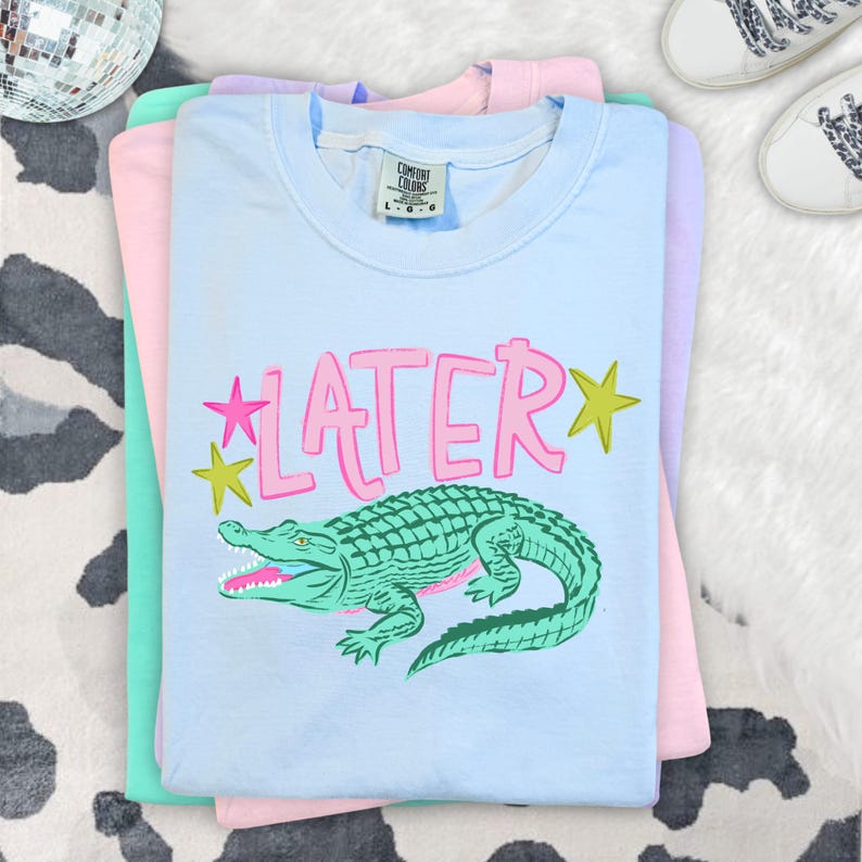 May include: Stacked pastel-colored t-shirts. The top shirt is light blue with a green alligator graphic and the word "LATER" in pink. The other shirts are pink and mint green. A disco ball and white sneakers are in the background.