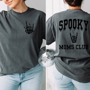 Pu&ograve; includere: Una maglietta grigio scuro con una mano scheletrica che fa un segno rock and roll sul davanti e il testo "SPOOKY MOMS CLUB" con un effetto gocciolante e una mano scheletrica che fa un segno rock and roll sul retro.