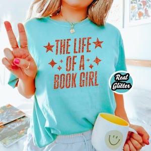 May include: A light blue t-shirt with the text "THE LIFE OF A BOOK GIRL" in glittery orange letters. The shirt features star and diamond accents. A white mug with a yellow interior and a smiley face is held in the hand.