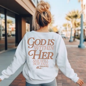 May include: White sweatshirt with the text "God is within her, she will not fall, Psalm 46:5" in brown and pink lettering. The text is in a vintage style font.