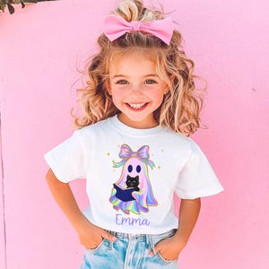 以下が含まれることがあります： ハロウィーンをテーマにしたグラフィックがプリントされた白いTシャツを着た少女。グラフィックは、黒猫を抱いた幽霊、虹色のリボン、紫色の筆記体で書かれた名前「Emma」が特徴です。少女は笑顔で、ピンクのリボンをしています。
