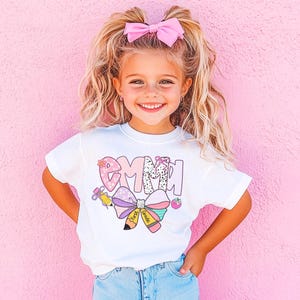 May include: A white t-shirt with the name "Emma" in a colorful, patterned design. The design includes a bow, pencils, and a strawberry. The girl in the image is wearing a pink bow in her hair and is smiling.