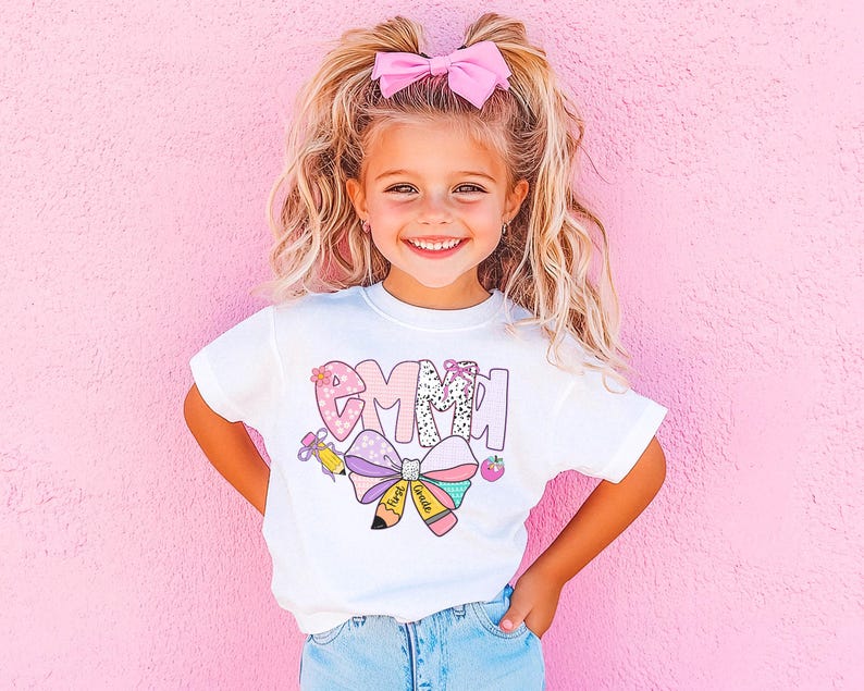 May include: A white t-shirt with the name "Emma" in a colorful, patterned design. The design includes a bow, pencils, and a strawberry. The girl in the image is wearing a pink bow in her hair and is smiling.