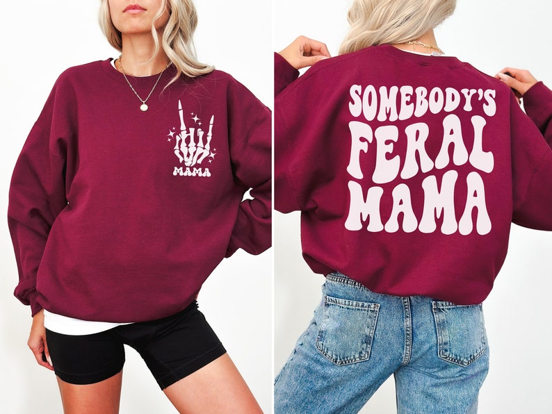 May include: Burgundy sweatshirt with white graphic text and design. The front features a skeleton hand making a rock-and-roll gesture with the word "MAMA" below. The back of the sweatshirt says "SOMEBODY'S FERAL MAMA" in a retro font.