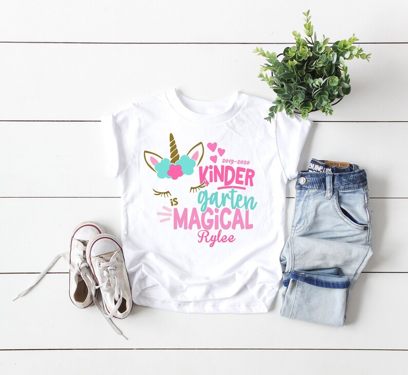 May include: White t-shirt with a unicorn graphic and the text "Kindergarten is Magical Rylee" in pink and teal. The unicorn has a gold horn, pink and teal flowers, and closed eyes. A pair of white sneakers and blue jeans are next to the shirt.