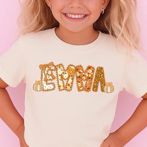 May include: A cream-colored t-shirt featuring the name "EMMA" in a floral and pumpkin-themed design. The letters are filled with various floral patterns in shades of orange, yellow, and white, with small pumpkin accents on each side.