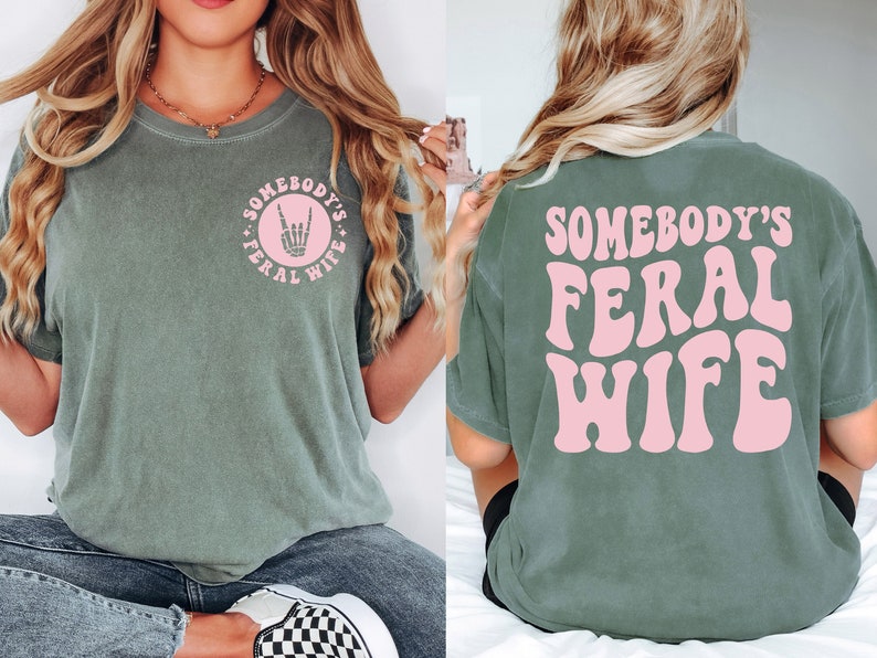 woman wearing a moss green comfort colors shirt with the pink wording on the back somebody's feral wife and a small pocket skeleton hand on the front