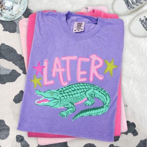 May include: A stack of three t-shirts in shades of purple and pink. The top shirt is purple with the word "LATER" in pink, along with a green alligator graphic. Two stars are on the left side of the word "LATER".