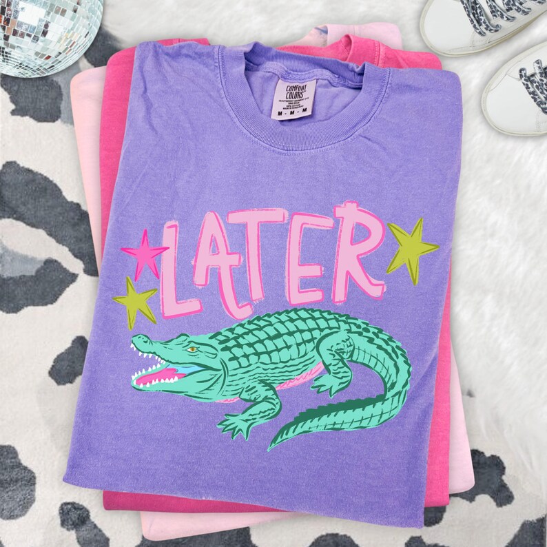 May include: A stack of three t-shirts in shades of purple and pink. The top shirt is purple with the word "LATER" in pink, along with a green alligator graphic. Two stars are on the left side of the word "LATER".