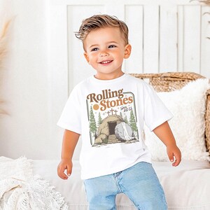 May include: A white t-shirt with the text "Rolling Stones Since 1933" and a graphic of a tomb, crosses, and trees. The shirt is worn by a young child in blue jeans.