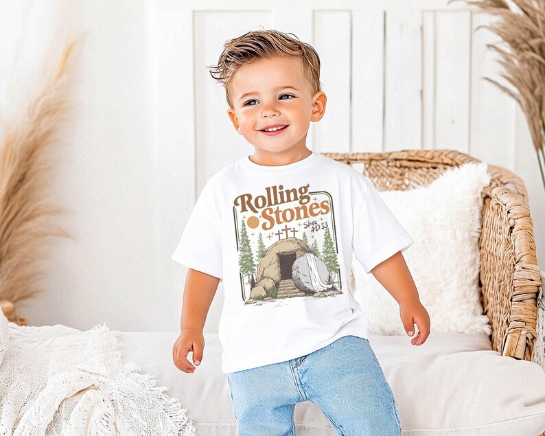 May include: A white t-shirt with the text "Rolling Stones Since 1933" and a graphic of a tomb, crosses, and trees. The shirt is worn by a young child in blue jeans.