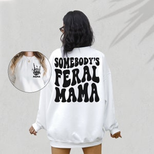 May include: White sweatshirt with the text "SOMEBODY'S FERAL MAMA" in black, retro-style lettering on the back. A smaller image on the front shows a skeleton hand making a rock-and-roll gesture, with the word "Mama" below.