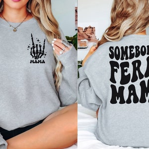 May include: A light gray sweatshirt with the text "SOMEBODY'S FERAL MAMA" on the back in black, retro-style lettering. The front features a black skeleton hand with the word "MAMA" below it. The sweatshirt is a casual style.