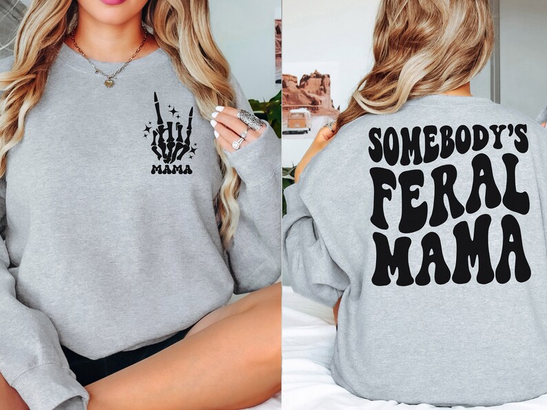 May include: A light gray sweatshirt with the text "SOMEBODY'S FERAL MAMA" on the back in black, retro-style lettering. The front features a black skeleton hand with the word "MAMA" below it. The sweatshirt is a casual style.